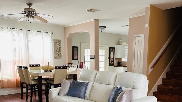 A spacious living room with a white leather sofa, a coffee table, and a dining area with a table and chairs. A staircase is visible in the background. - Spacious 4BR 2-Story North Houston Vacation Rental (Tomball)