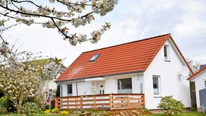 Een wit huis met een rood dak, een houten omheining en een tuin vol bloemen.