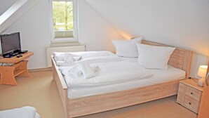 Een net opgemaakt bed in een kamer met een schuine balkenkap, een klein bureau met een computer en een bedlampje.