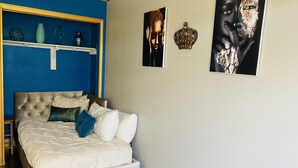 A bedroom with a tufted headboard, a white bedspread, and decorative pillows. There are wall-mounted shelves with glass vases and a crown-shaped ornament. - Spacious North Houston Getaway rental (Tomball)