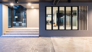 Entrada de un edificio moderno con puerta de cristal, escalones de mármol y un amplio ventanal con persianas verticales.