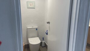 A small, white bathroom with a toilet, a blue cleaning brush, and a framed picture on the wall. - Oceanic Apartments Unit 13 Stunning Ocean Views (Kings Beach)