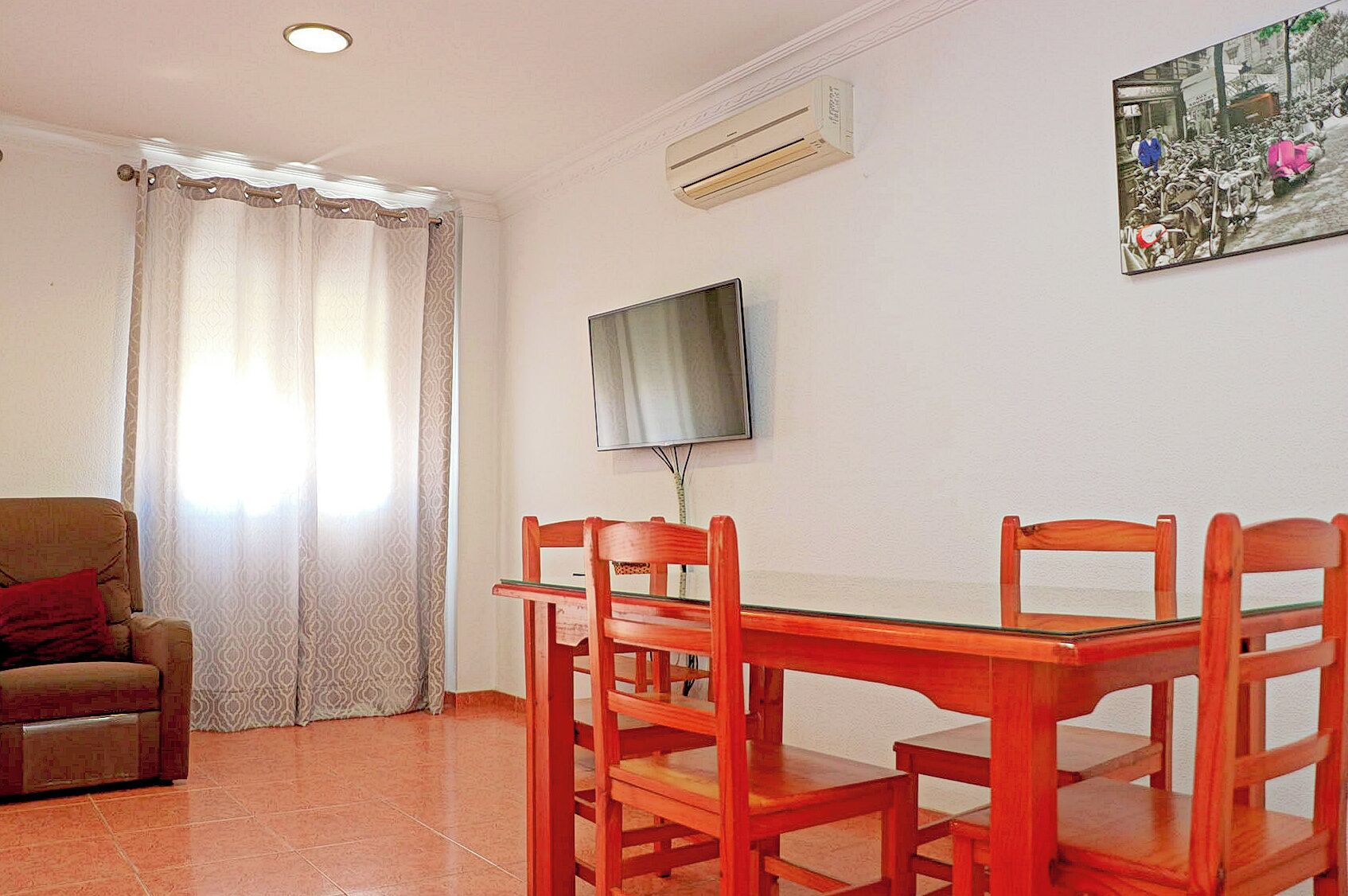 A living room with a sofa, a dining table with chairs, a TV mounted on the wall, and a patterned curtain.