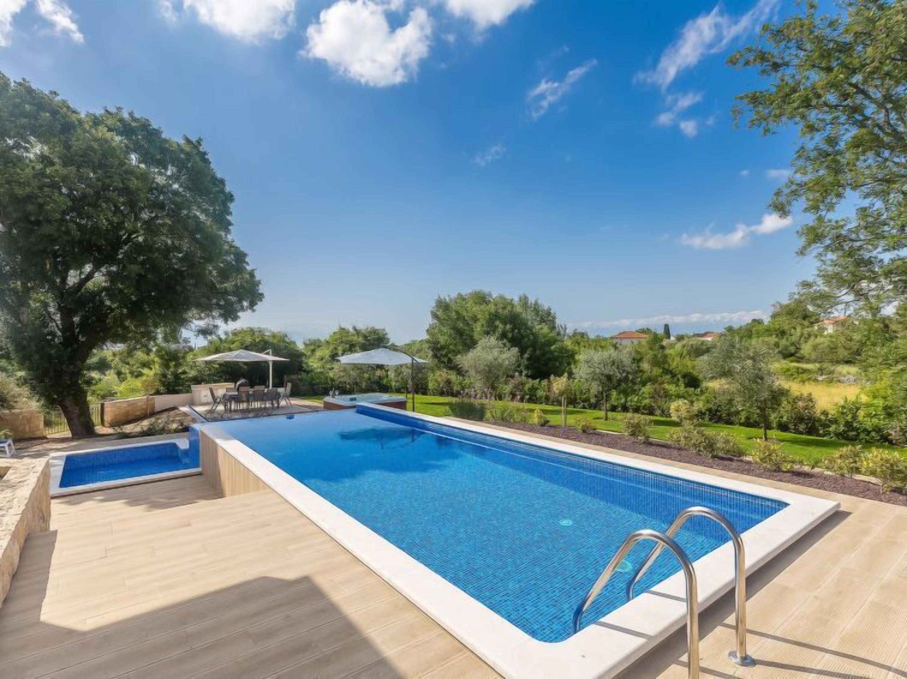 A rectangular swimming pool with a wooden deck, surrounded by lush greenery and trees.