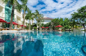 A swimming pool with red umbrellas, palm trees, and a building with balconies.