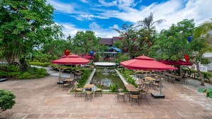 An outdoor patio area with tables and chairs under red umbrellas, surrounded by lush greenery and a small pond. - Chez Carole Center Resort & Spa (Phu Quoc)