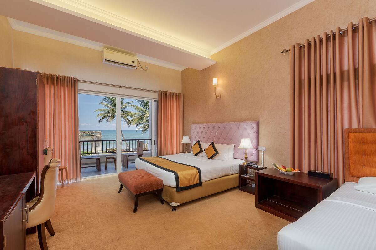 A hotel room with a large bed, a desk, a chair, and a balcony with a view of palm trees.