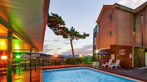 A swimming pool with lounge chairs and a deck area, surrounded by a brick building and a view of the sunset. - Goorm Dari Pension (Incheon)