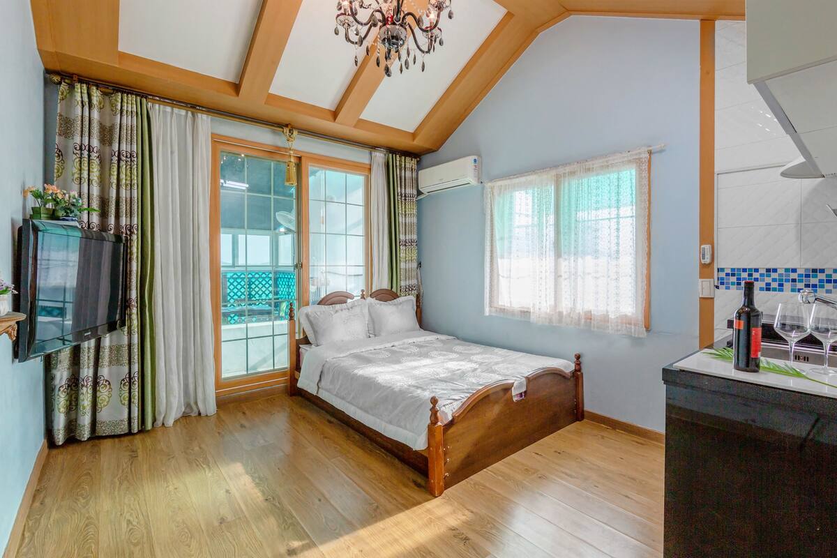 A bedroom with a wooden bed, a TV, a balcony, and a kitchen area.