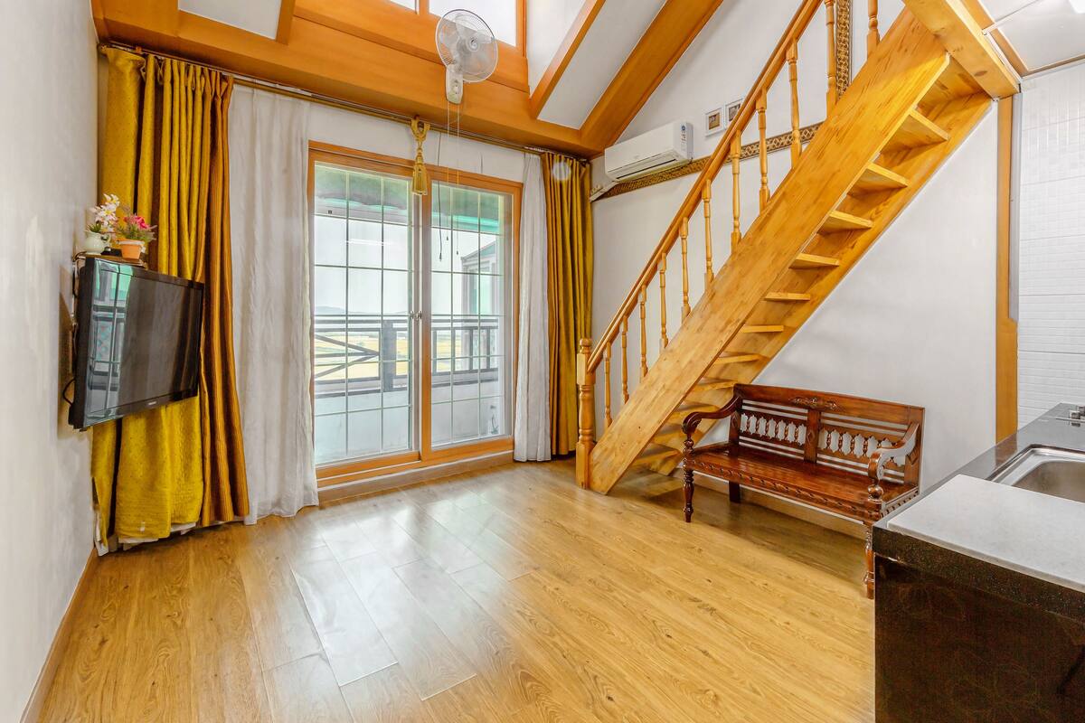 A spacious room with a wooden staircase, a bench, a kitchen area, and large windows with curtains.