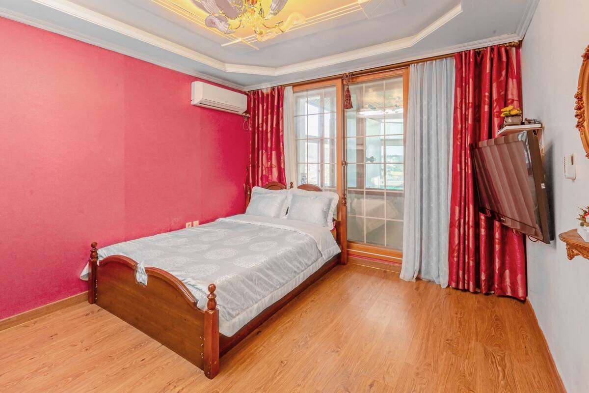 A bedroom with a wooden bed, a large window with curtains, an air conditioning unit, and a television on the wall.