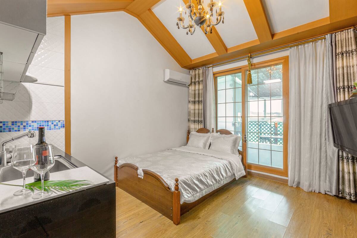 A bedroom with a bed, a window with curtains, a TV, and a bathroom with a sink and a bottle.