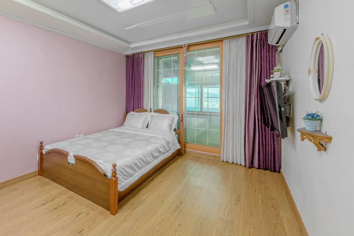 A bedroom with a wooden bed, a large window with curtains, an air conditioning unit, and a small shelf with a potted plant.