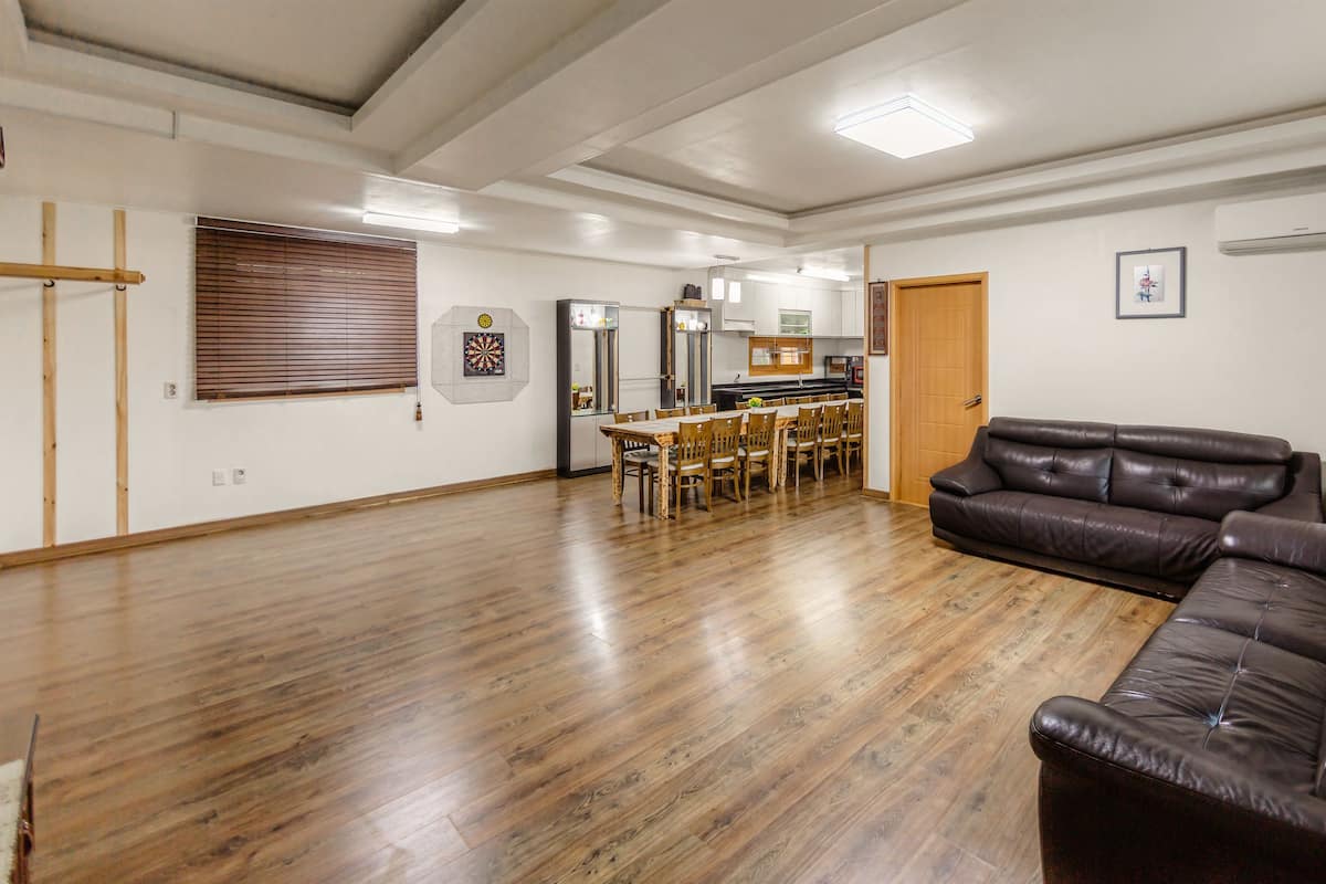 A spacious living room with a wooden floor, a leather sofa, a dining table with chairs, and a kitchen area with cabinets and appliances.
