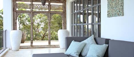 A modern living room with a grey sofa, patterned cushions, a hanging lantern, and a large window with a view of trees.