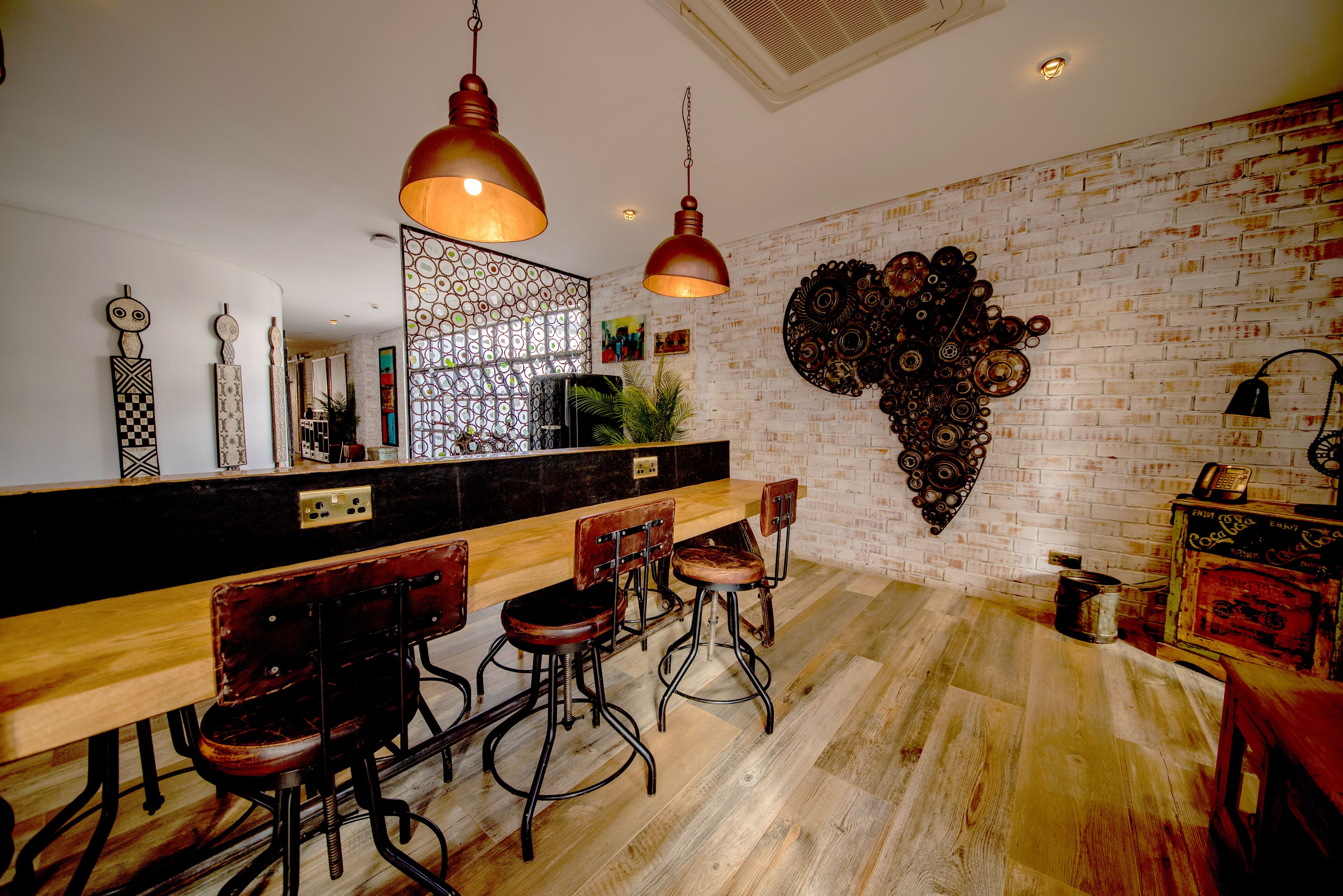 A modern kitchen with a long wooden bar, bar stools, hanging pendant lights, and a brick wall with decorative metal art.