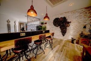 A modern kitchen with a long wooden bar, bar stools, hanging pendant lights, and a brick wall with decorative metal art.