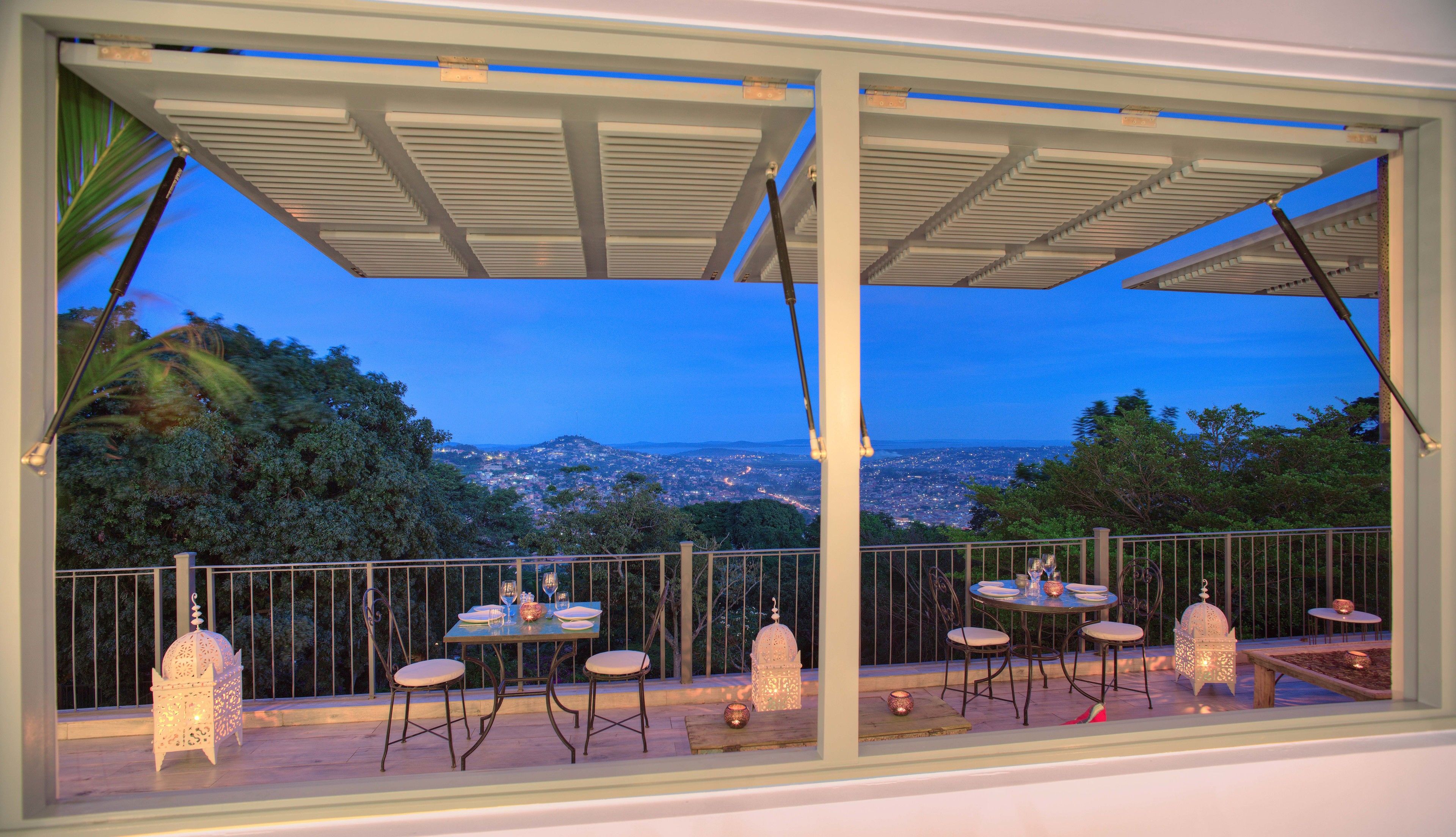 An outdoor dining area with tables and chairs, decorative lanterns, and a view of a cityscape at dusk.