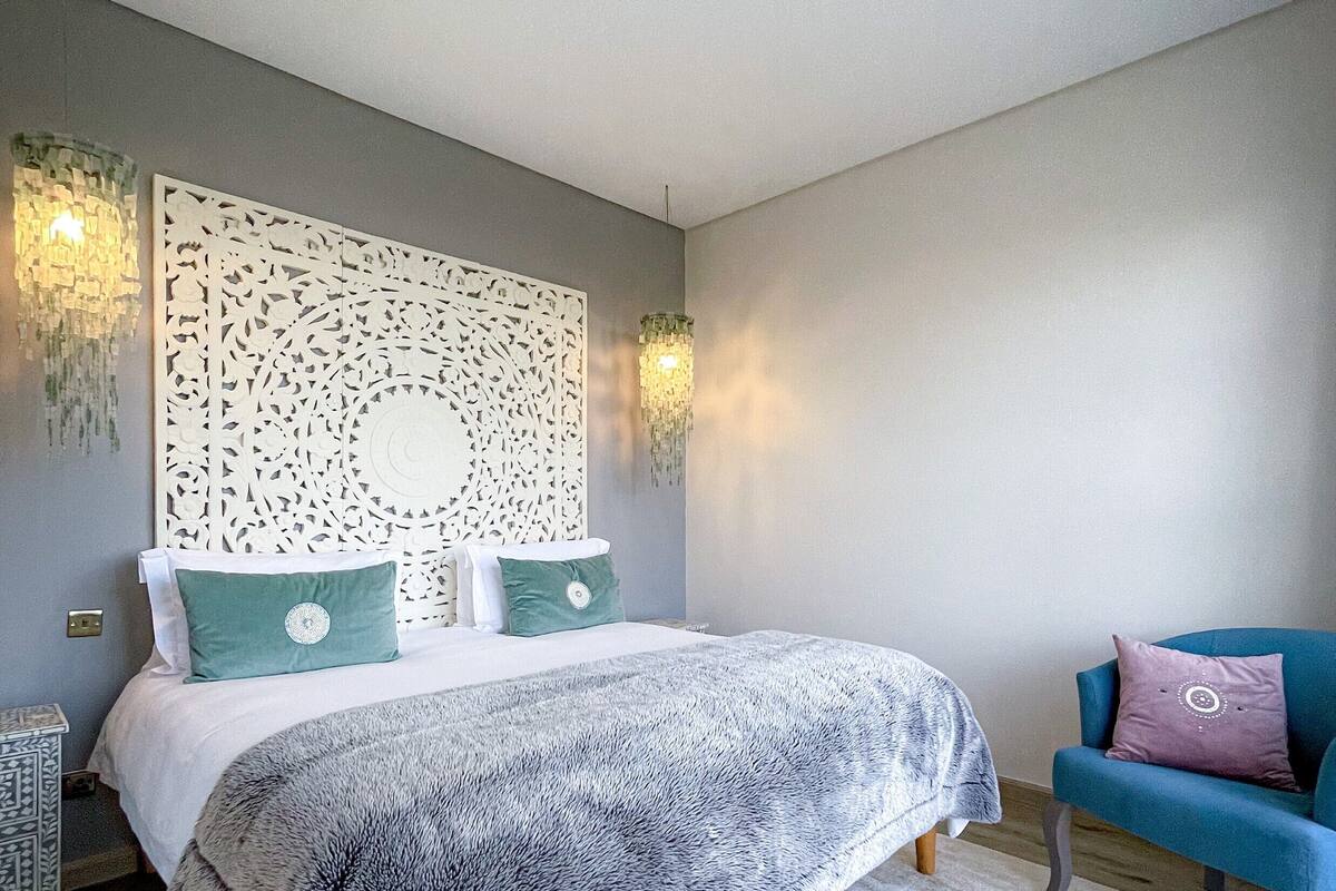 A bedroom with a large bed, a decorative headboard, a blue armchair, and a patterned rug.