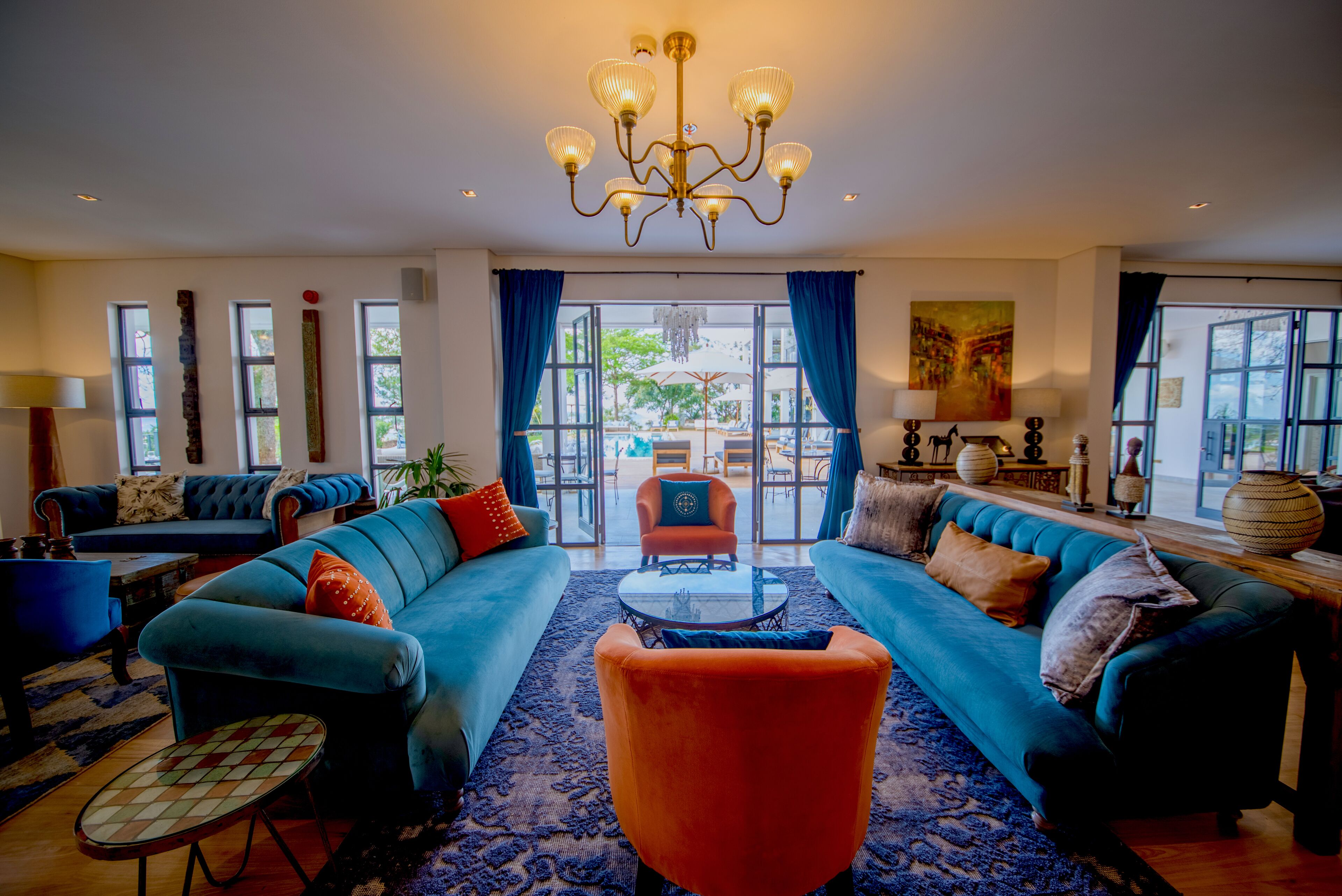 A spacious living room with a large blue sofa, a smaller orange armchair, and a coffee table. There is a chandelier, a painting on the wall, and a view through large windows.