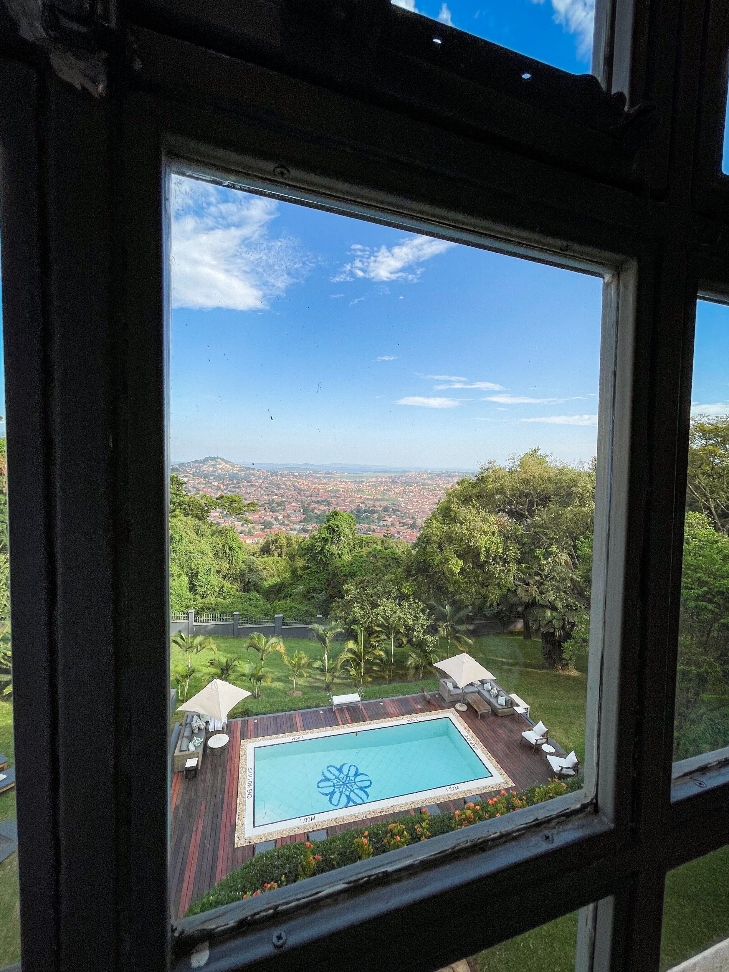 Vista desde una ventana a una piscina con diseño geométrico, rodeada por una terraza y sillas de descanso, con un paisaje urbano al fondo.