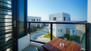 Un balcón con una mesa de madera y un ventanal de vidrio que da a un patio con edificios modernos y una vista despejada del mar.