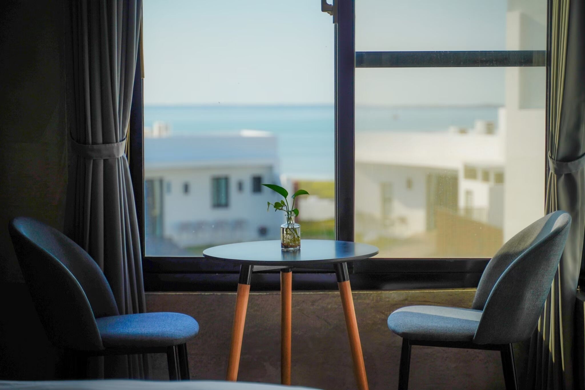 A room with a view of the sea, two chairs, a small table with a vase, and a window with curtains.