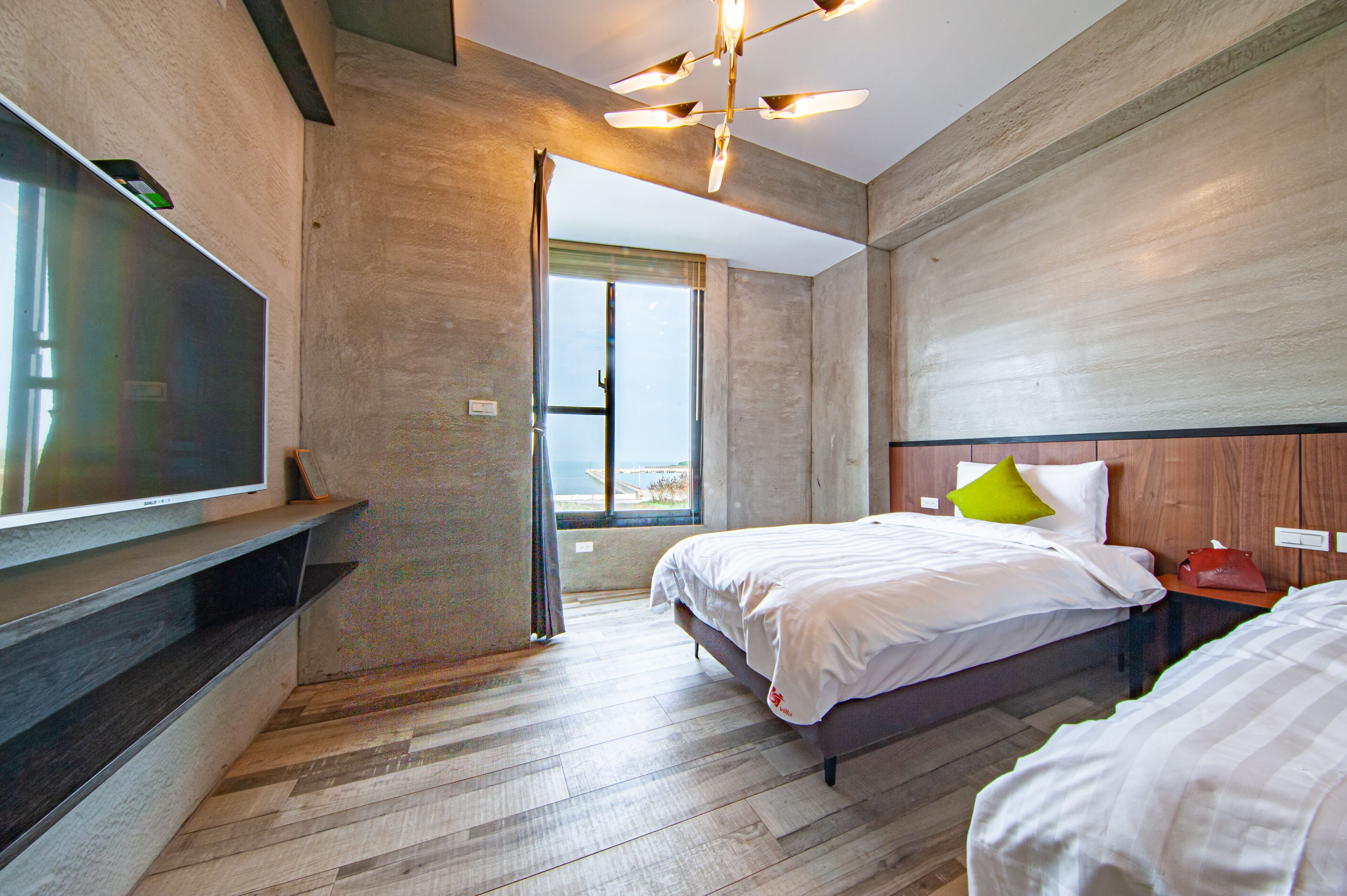 A modern hotel room with a large flat-screen TV mounted on the wall, a bed with white bedding and a green pillow, and a wooden headboard.