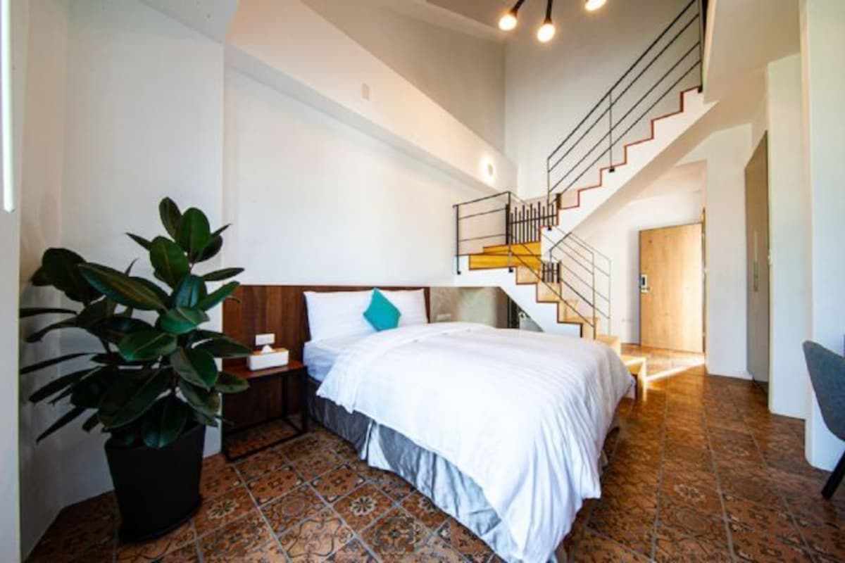 Un dormitorio con cama, mesita de noche, planta en maceta y una escalera que lleva a otra habitación.