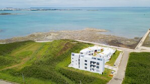 Un moderno edificio blanco de varios pisos y balcones, ubicado en una ladera cubierta de césped con vistas a una zona costera con costas rocosas y aguas azules tranquilas.