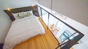 A bedroom with a wooden floor, a bed with white bedding, and a large window.