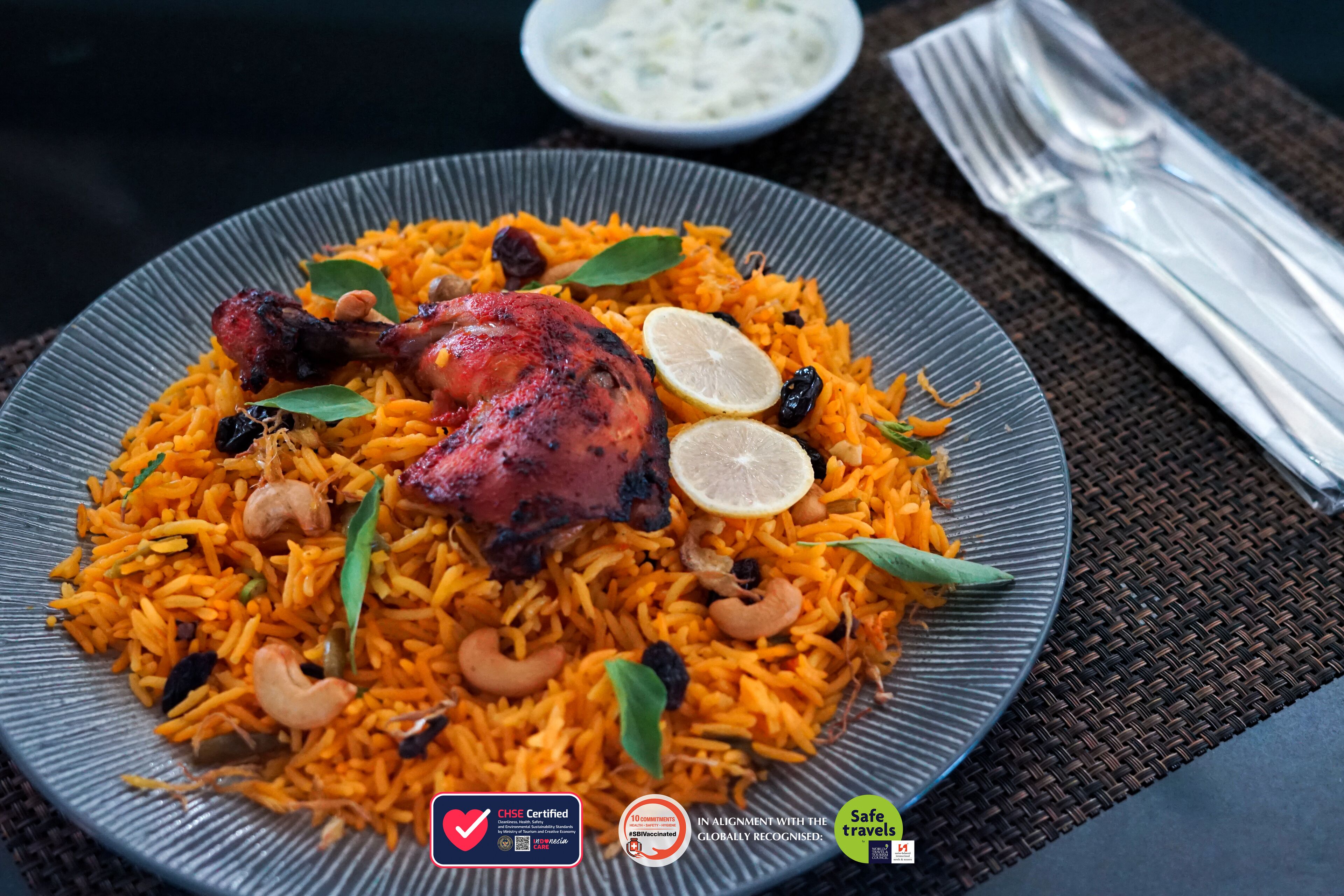 Um prato de biryani com um pedaço de frango assado, decorado com rodelas de limão e nozes, sobre um prato texturizado.