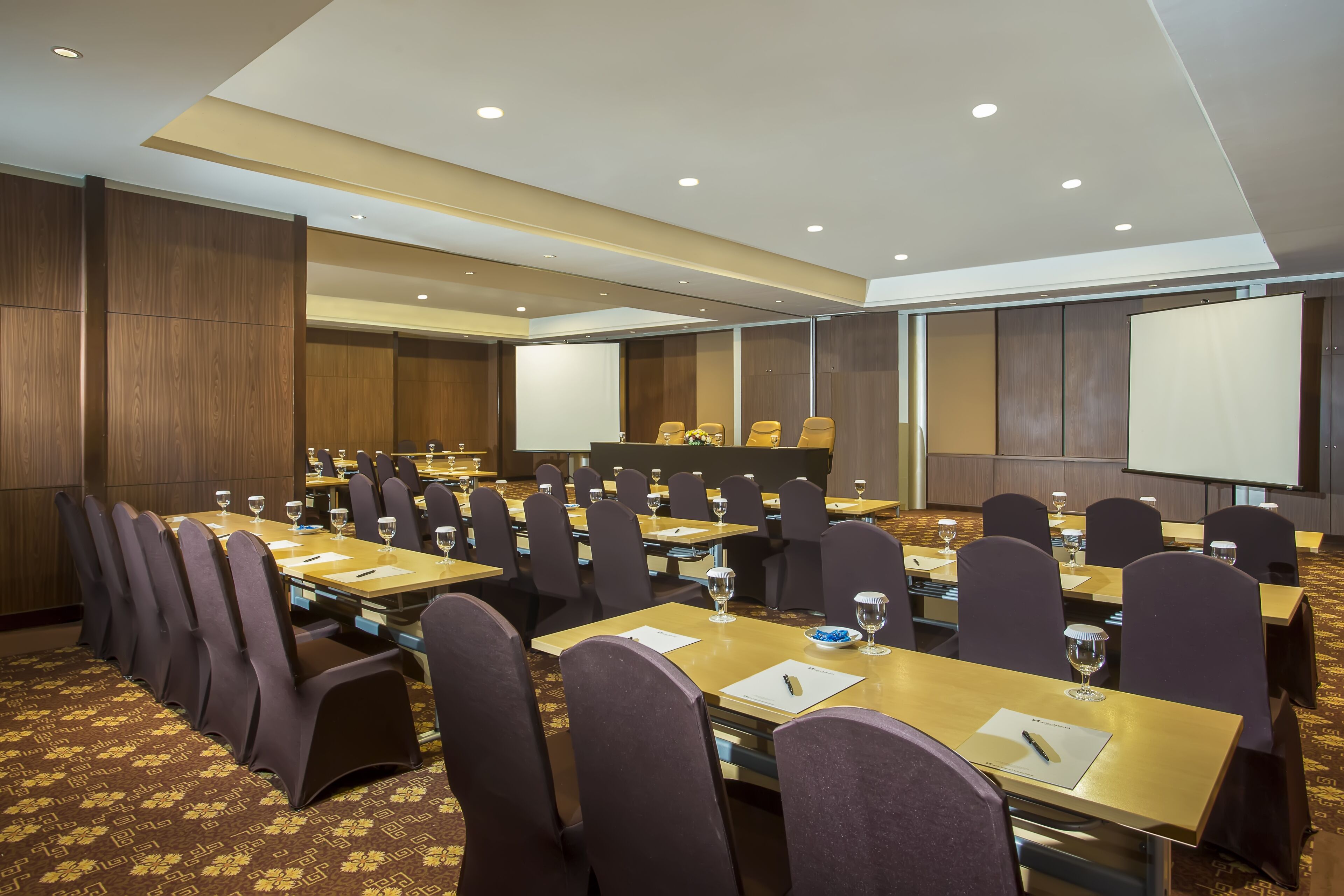 A conference room with multiple tables and chairs, equipped with nameplates, water glasses, and a projector screen.