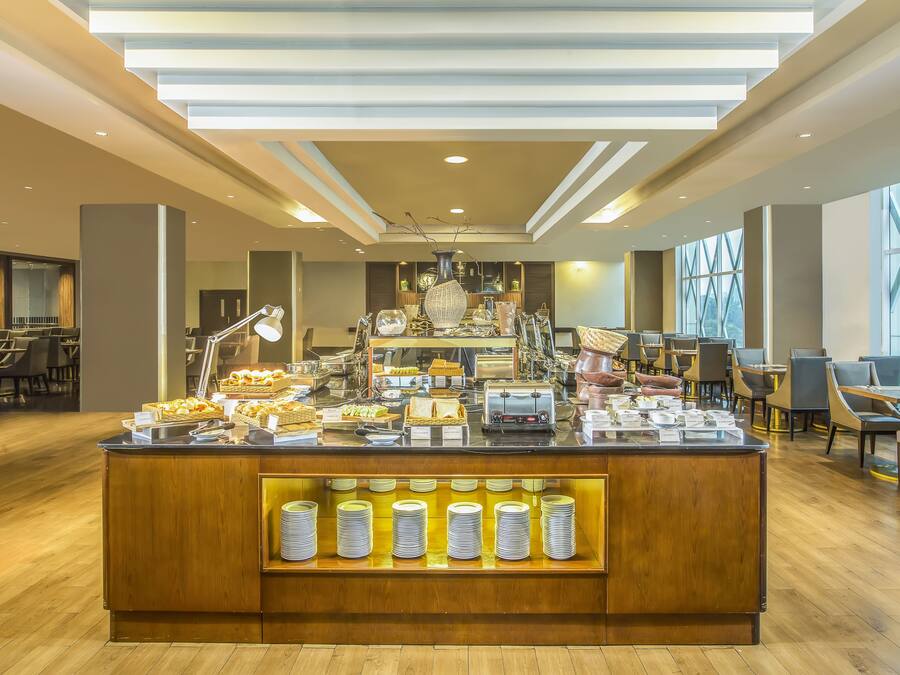 Zona de buffet con una variedad de alimentos, una barra con platos y un área de comedor con mesas y sillas.