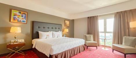 A hotel room with a large bed, two chairs, a nightstand with a lamp, and a window with curtains.