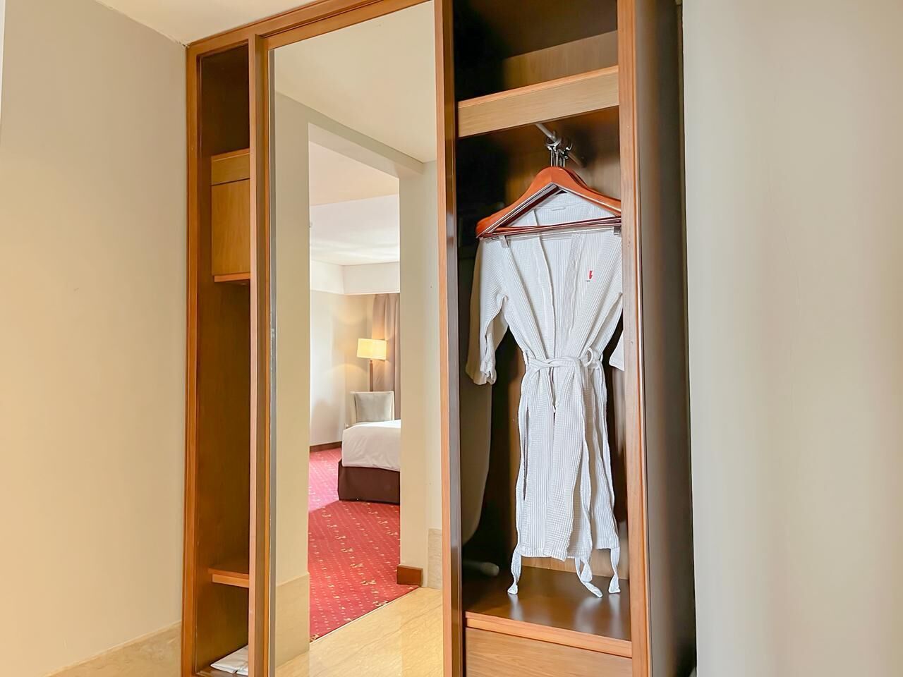 A hotel room with a wooden wardrobe, a hanging robe, and a bed with a red carpet.