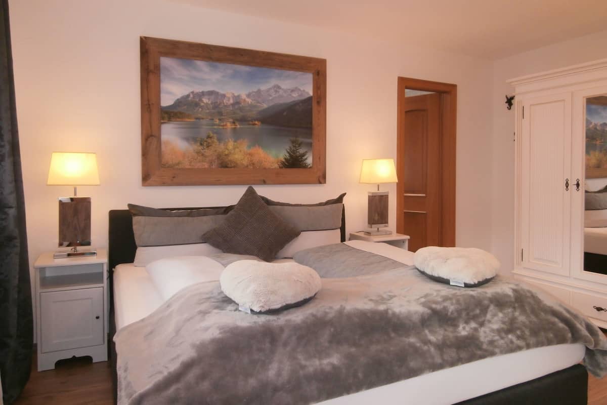 A bedroom with a bed, two bedside tables, a nightstand, a painting of a mountain landscape, and a wardrobe.