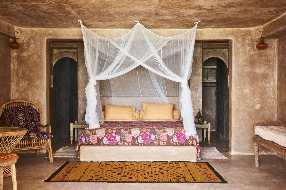 A room with a four-poster bed, a patterned bedspread, a woven rug, and a wicker chair.