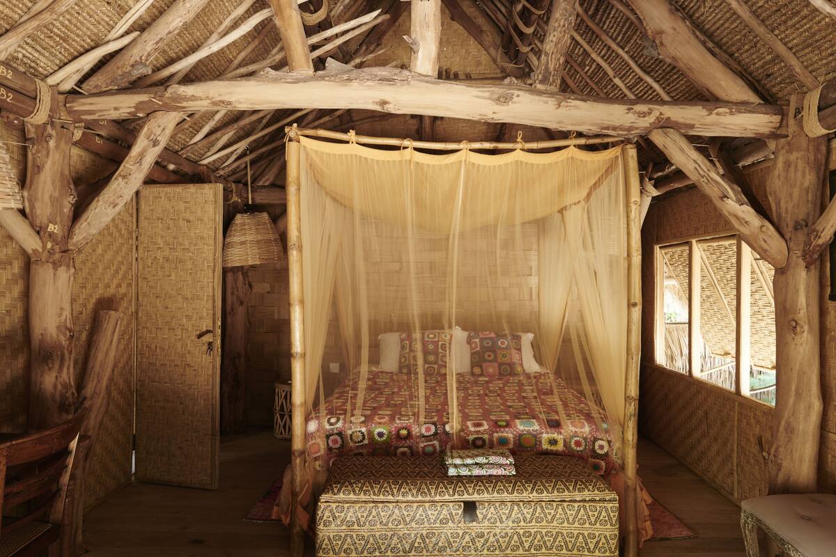 A rustic wooden room with a bed under a canopy, a woven basket, and a wooden door.