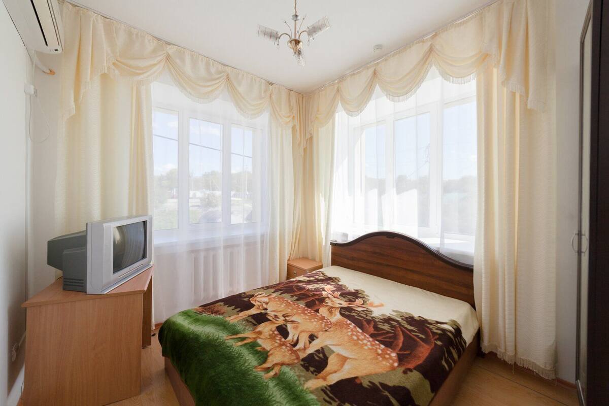 Un dormitorio con cama, televisor sobre un mueble de madera y ventanas amplias con cortinas translúcidas.