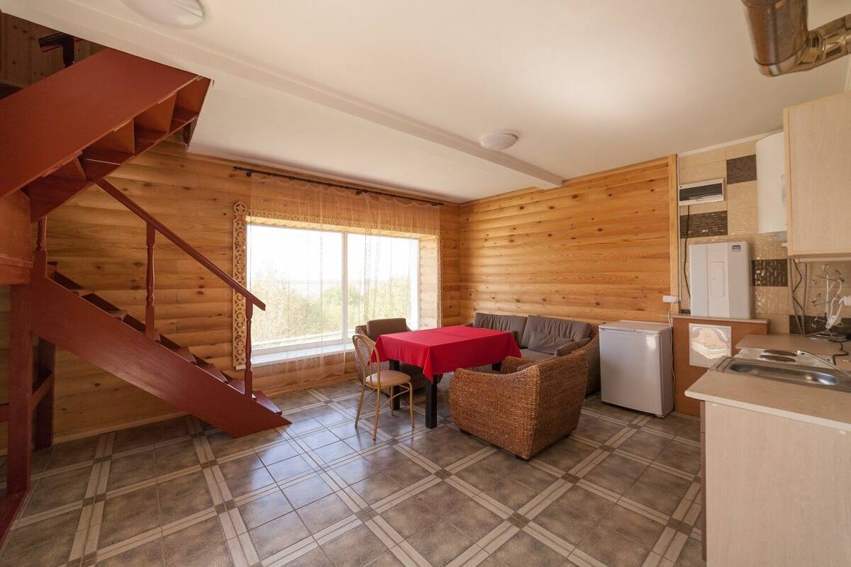 Interior de una cabaña de troncos con una escalera, un comedor con mesa roja, muebles de mimbre y una kitchenette.