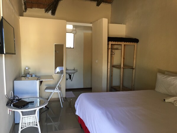 A hotel room with a bed, a desk with a chair, a small table, a mirror, a sink, and a wardrobe. - Athoma Guest House (Shakawe)