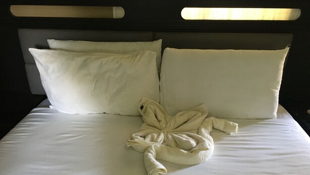 Una cama bien hecha con sábanas y almohadas blancas, una bata doblada sobre la cama y un cabecero con una lámpara.
