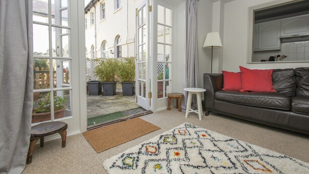 A living room with a sofa, a patterned rug, a small stool, a lamp, and a view through a glass door to an outdoor area with potted plants.