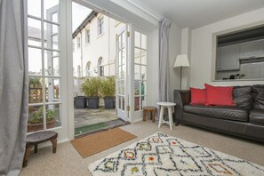 Een woonkamer met een bank, een vloerkleed met patroon, een klein krukje, een lamp, en uitzicht door een glazen deur op een buitenruimte met plantenpotten.