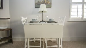 A dining table with white chairs, two wine glasses, and a table lamp. There is a painting on the wall and a window with blinds.