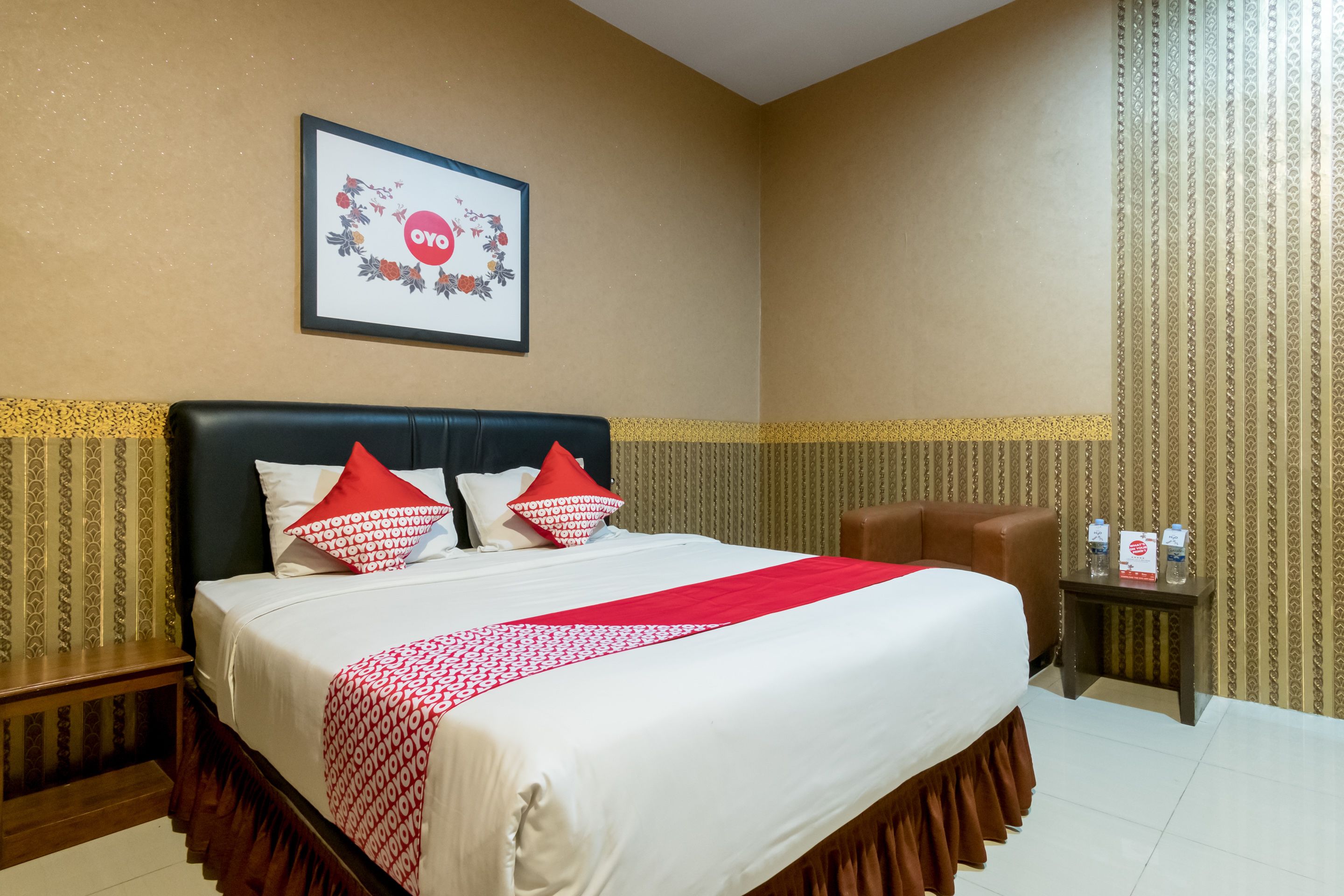 A hotel room with a bed, a framed picture on the wall, a small table with water bottles, and a chair.