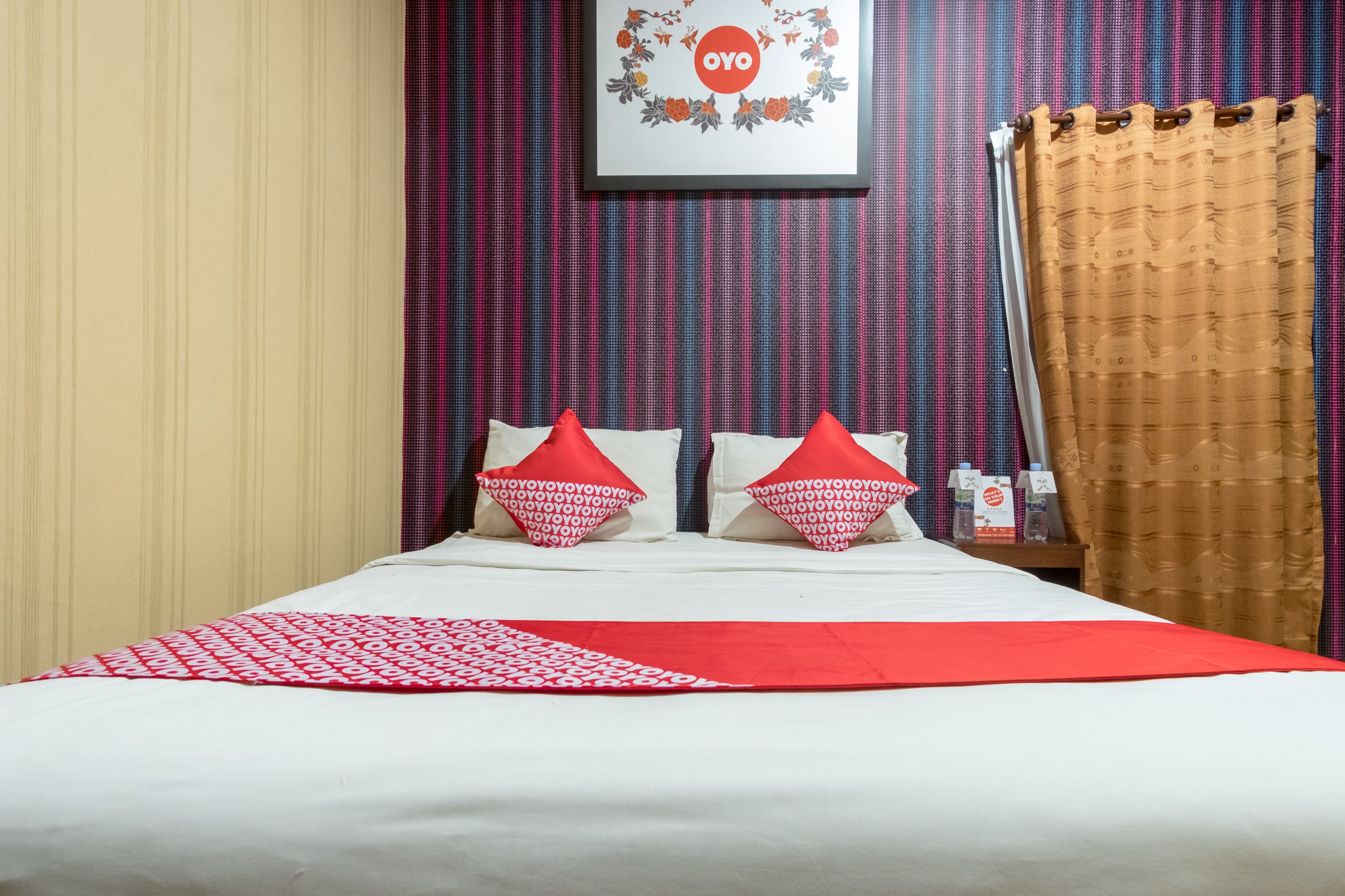 A hotel room with a bed, red and white pillows, a red throw blanket, a framed picture on the wall, and a curtain.