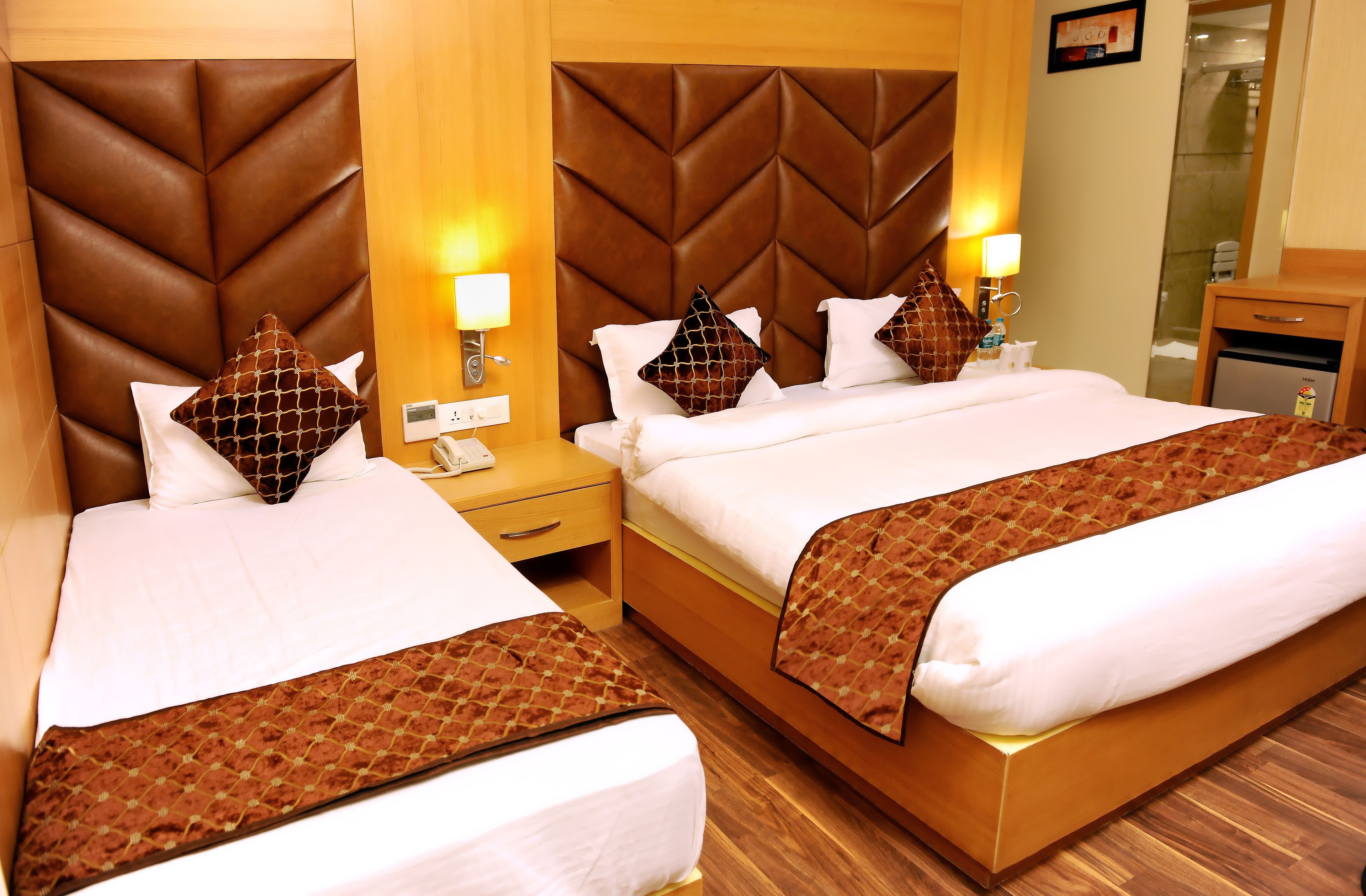 A hotel room with two beds, each with a brown headboard and white bedding. There are bedside lamps, a nightstand, and a small table with a lamp.