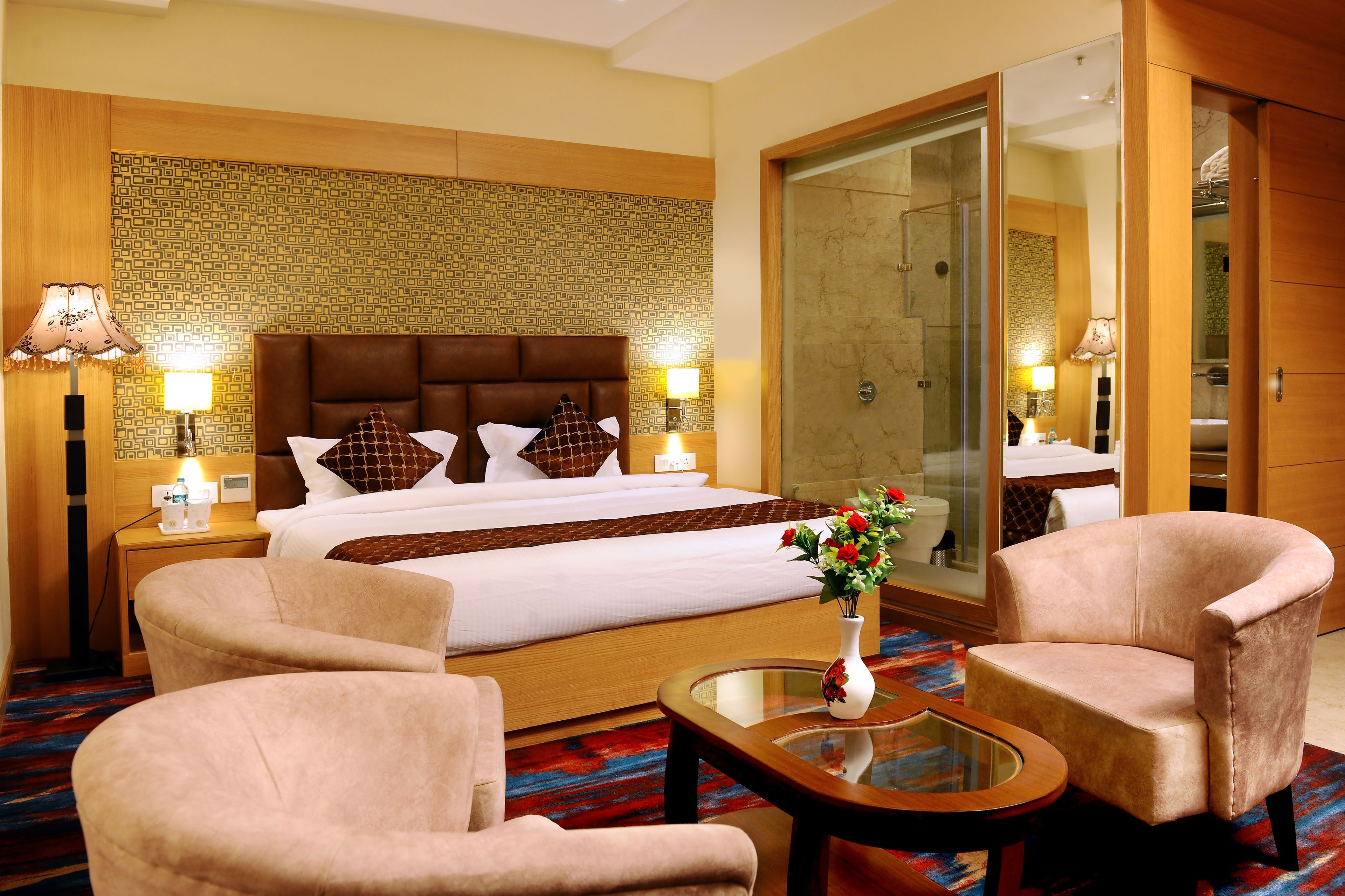 A hotel room with a large bed, two bedside tables with lamps, a glass coffee table, two armchairs, and a mirror.